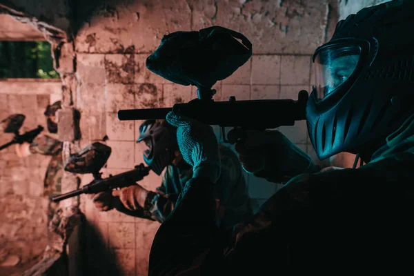 Vista de perto da equipe de paintball em máscaras uniformes e protetoras jogando paintball com armas de marcador em prédio abandonado — Fotografia de Stock