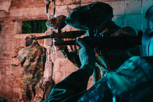 Partial view of paintball team in uniform and protective masks playing paintball with marker guns in abandoned building — Stock Photo