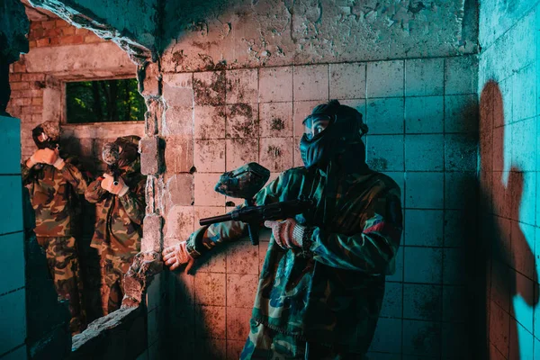 Male paintball player in goggle mask and camouflage uniform hiding behind wall while other team is standing near in abandoned building — Stock Photo