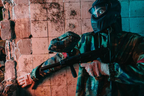 Male paintball player in camouflage and goggle mask hiding behind wall and holding marker gun in abandoned building — Stock Photo