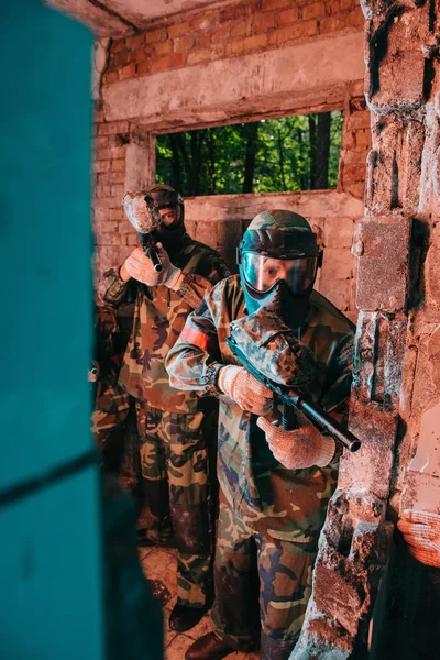 Пейнтбольная команда в форме и защитных масках, играющая с пейнтбольными пушками в заброшенном здании — стоковое фото