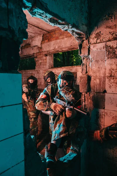 Equipo de paintball en uniforme y máscaras protectoras jugando paintball con pistolas marcadoras en edificio abandonado - foto de stock