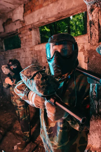 Jugador de paintball masculino con máscara de gafas y uniforme de camuflaje de pie mientras otro jugador apuntaba pistola de paintball en su cuello en un edificio abandonado - foto de stock