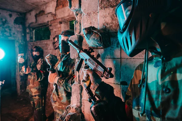 Vista parcial del jugador de paintball masculino con máscara de gafas y uniforme de camuflaje escondido detrás de la pared y apuntando pistola marcadora al jugador de otro equipo en edificio abandonado - foto de stock