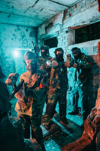 Paintball team in uniform and protective masks aiming by paintball guns at camera in abandoned building — Stock Photo