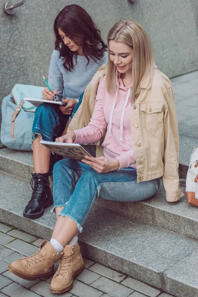 Mujeres jóvenes con cuadernos descansando en los escalones de la ciudad durante el viaje - foto de stock