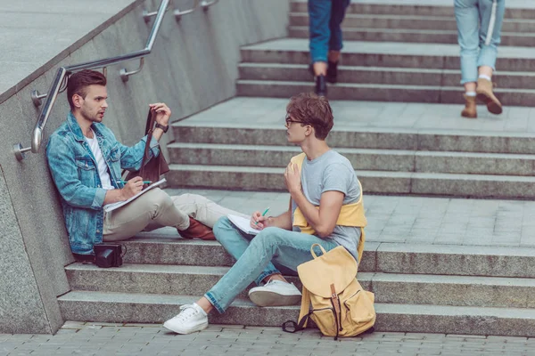 Homens jovens com cadernos sentados em degraus da cidade em nova cidade — Fotografia de Stock
