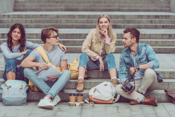 Young tourists with backpacks and coffee to go resting on steps on street of new city — Stock Photo