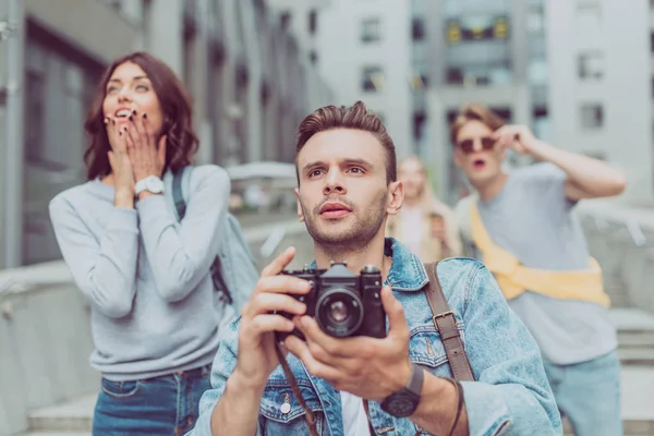 Selective focus of tourist with photo camera in hands and shocked friends on street behind — Stock Photo