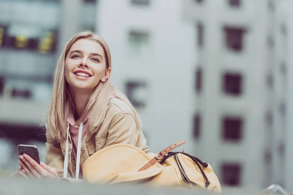 Happy young blonde woman with backpack using smartphone in city — Stock Photo