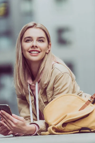 Усміхнена блондинка з рюкзаком за допомогою смартфона — стокове фото