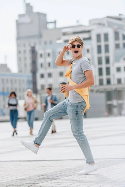 Excited tourist with coffee to go and backpack walking in city with friends behind — Stock Photo