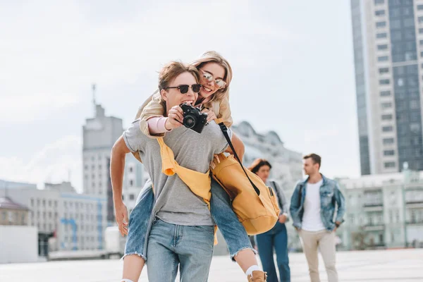 Feliz namorado dando piggyback para namorada com câmera de fotos na cidade com amigos atrás — Fotografia de Stock