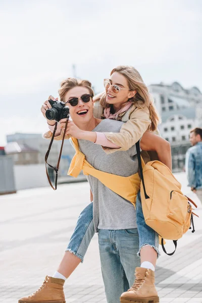 Happy boyfriend giving piggyback to girlfriend with photo camera in city — Stock Photo