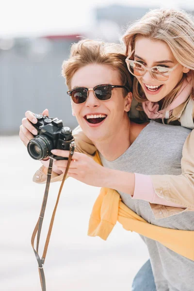 Smiling tourist giving piggyback to happy girlfriend while she taking photo on camera — Stock Photo