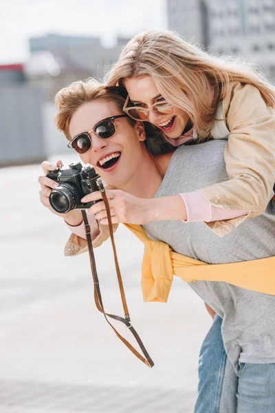 Petit ami souriant donnant piggyback à belle petite amie pendant qu'elle prend une photo sur la caméra — Photo de stock