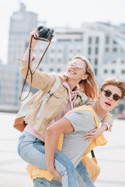 Boyfriend piggybacking smiling girlfriend while she taking photo on camera — Stock Photo