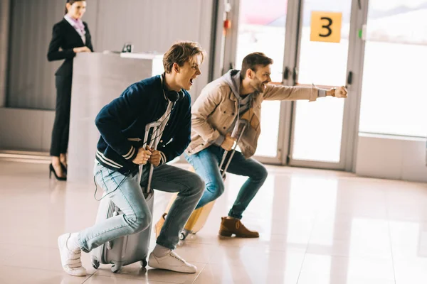 Fröhliche junge Männer, die Spaß haben und Koffer fahren im Flughafen — Stockfoto