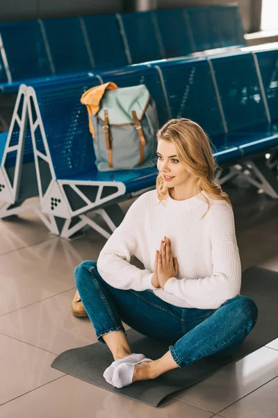 Blick aus der Vogelperspektive auf eine lächelnde junge Frau, die in Lotusposition meditiert, während sie im Flughafenterminal auf ihren Flug wartet — Stockfoto