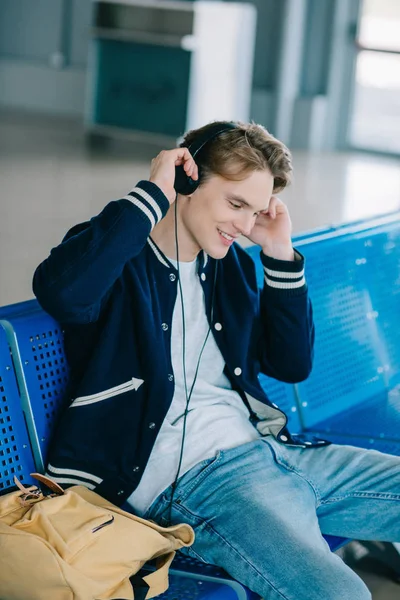 Lächelnder junger Mann mit Kopfhörern, der am Flughafen sitzt und auf seinen Flug wartet — Stockfoto