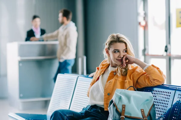 Joven sonriente con mochila sentada y mirando hacia otro lado en la terminal del aeropuerto - foto de stock