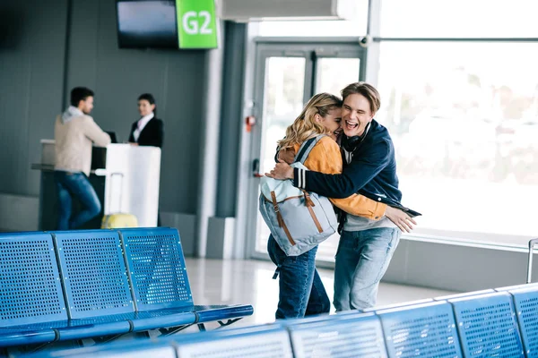 Збуджена молода пара з рюкзаками, що обіймаються в терміналі аеропорту — стокове фото