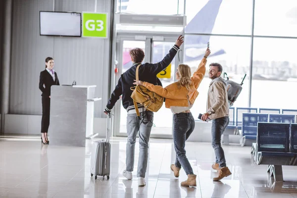Visão traseira de jovens amigos felizes acenando as mãos e indo para a mesa de check-in no aeroporto — Fotografia de Stock