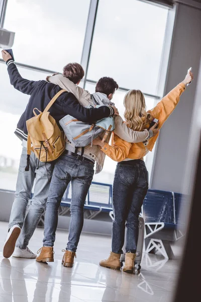 Назад вид счастливых молодых друзей с паспортами обнимающих и поднимающих руки в аэропорту — стоковое фото