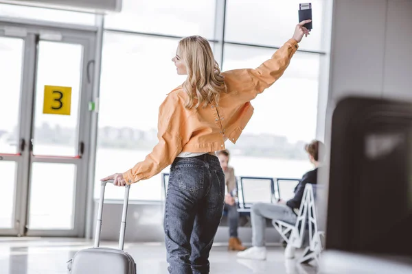 Back view of happy young woman holding passport and suitcase in airport — Stock Photo