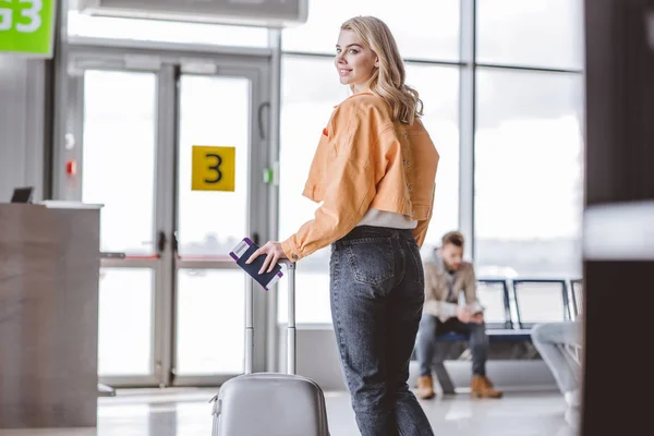 Girl with passport, boarding pass and suitcase smiling at camera in airport — Stock Photo