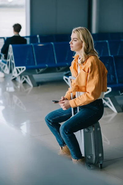 Enfoque selectivo de la mujer joven con pasaporte sentado en la maleta y mirando hacia otro lado en el aeropuerto — Stock Photo