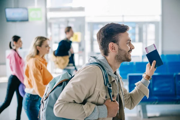 Вид сбоку улыбающегося человека с рюкзаком в руках с паспортом и посадочным талоном в аэропорту — стоковое фото