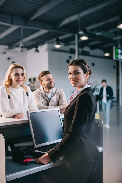 Young airport worker checking documents and smiling at camera at check-in desk in airport — Stock Photo