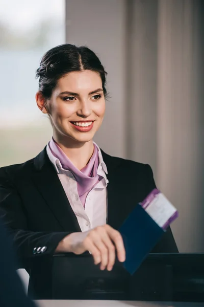 Beautiful smiling young airport worker holding passport with boarding pass at check-in desk — Stock Photo