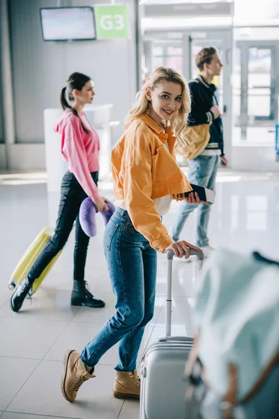Chica atractiva con la maleta sonriendo a la cámara en el aeropuerto - foto de stock