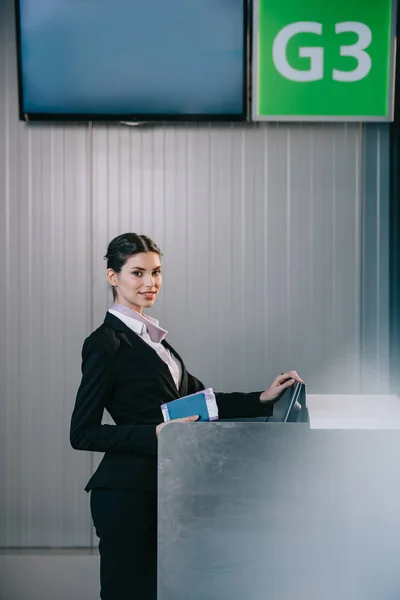 Hermosa joven trabajador sosteniendo documentos y sonriendo a la cámara en el mostrador de facturación en el aeropuerto - foto de stock