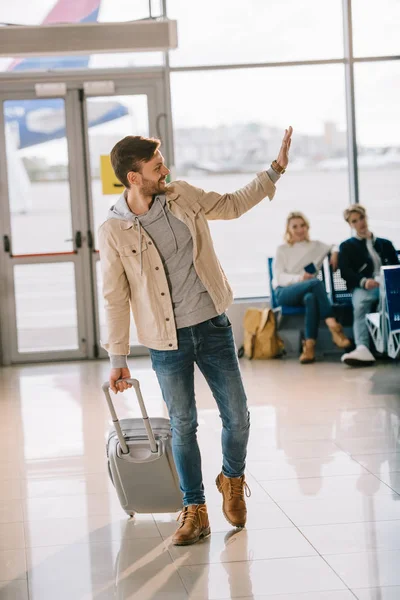 Щасливі молода людина з чемодан розмахуючи рукою і фотографіях хтось дивитися вбік в аеропорту — стокове фото