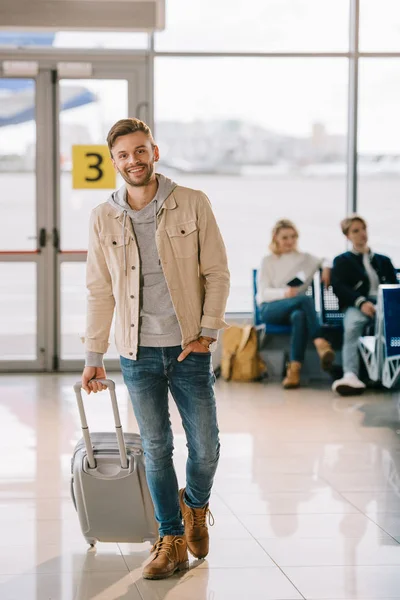 Joven guapo con maleta sonriendo a la cámara en el aeropuerto - foto de stock