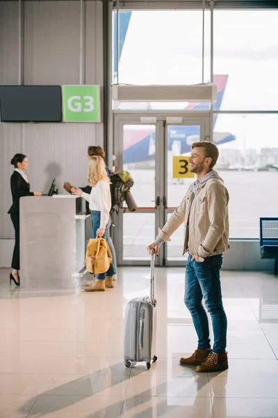 Vista lateral do jovem sorridente de pé com mala no aeroporto — Stock Photo