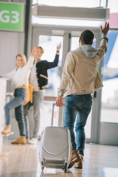 Вид ззаду чоловіка з валізою махає рукою друзям на стійці реєстрації в аеропорту — стокове фото