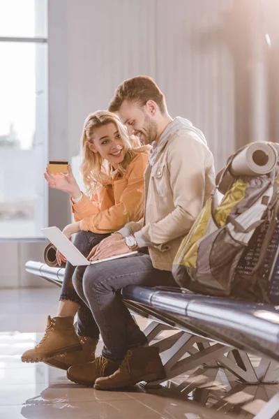 Sonriente chica celebración de la tarjeta de crédito, mientras que el hombre usando ordenador portátil en la terminal del aeropuerto — Stock Photo