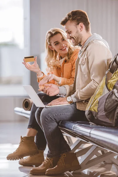 Sonriente joven que muestra la tarjeta de crédito al hombre feliz utilizando el ordenador portátil en el aeropuerto - foto de stock