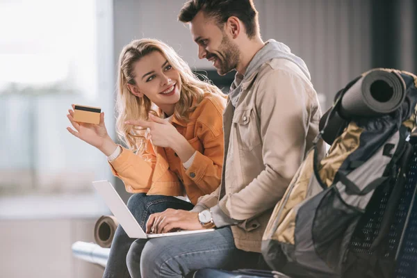 Smiling girl pointing with finger at credit card and looking at man using laptop in airport — Stock Photo