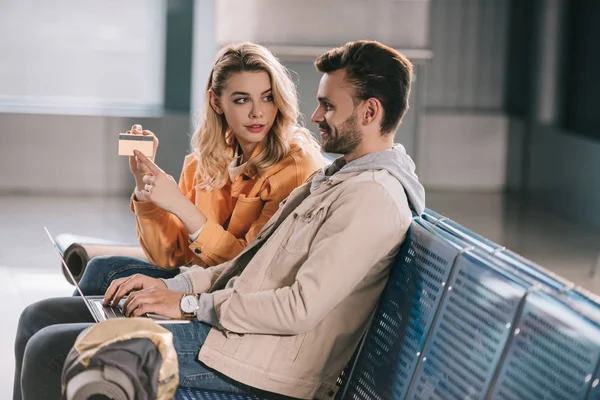 Chica mostrando tarjeta de crédito al hombre sonriente utilizando el ordenador portátil en el aeropuerto - foto de stock