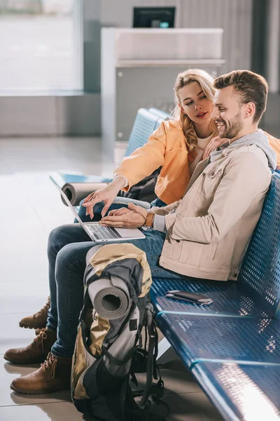 Chica mirando sonriente hombre usando el ordenador portátil mientras está sentado en la terminal del aeropuerto - foto de stock