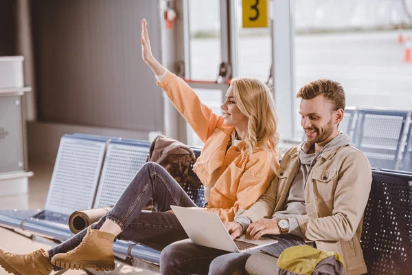 Joven sonriente usando el ordenador portátil y la mujer saludando de la mano mientras están sentados y esperando juntos en el aeropuerto - foto de stock
