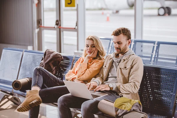 Jeune couple de voyageurs utilisant un ordinateur portable et attendant le vol dans le terminal de l'aéroport — Photo de stock