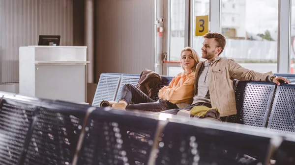 Messa a fuoco selettiva di giovani coppie con zaini seduti insieme e in attesa di volo in aeroporto — Foto stock