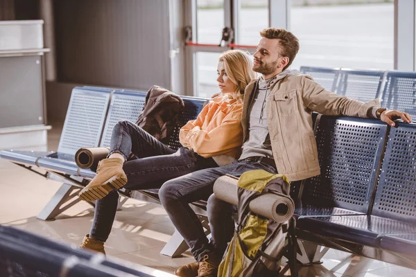 Pareja joven con mochilas sentadas juntas y esperando el vuelo en el aeropuerto - foto de stock