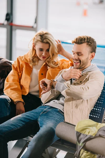 Junges Paar sitzt am Flughafen und checkt Armbanduhr, während es auf den Flug wartet — Stockfoto